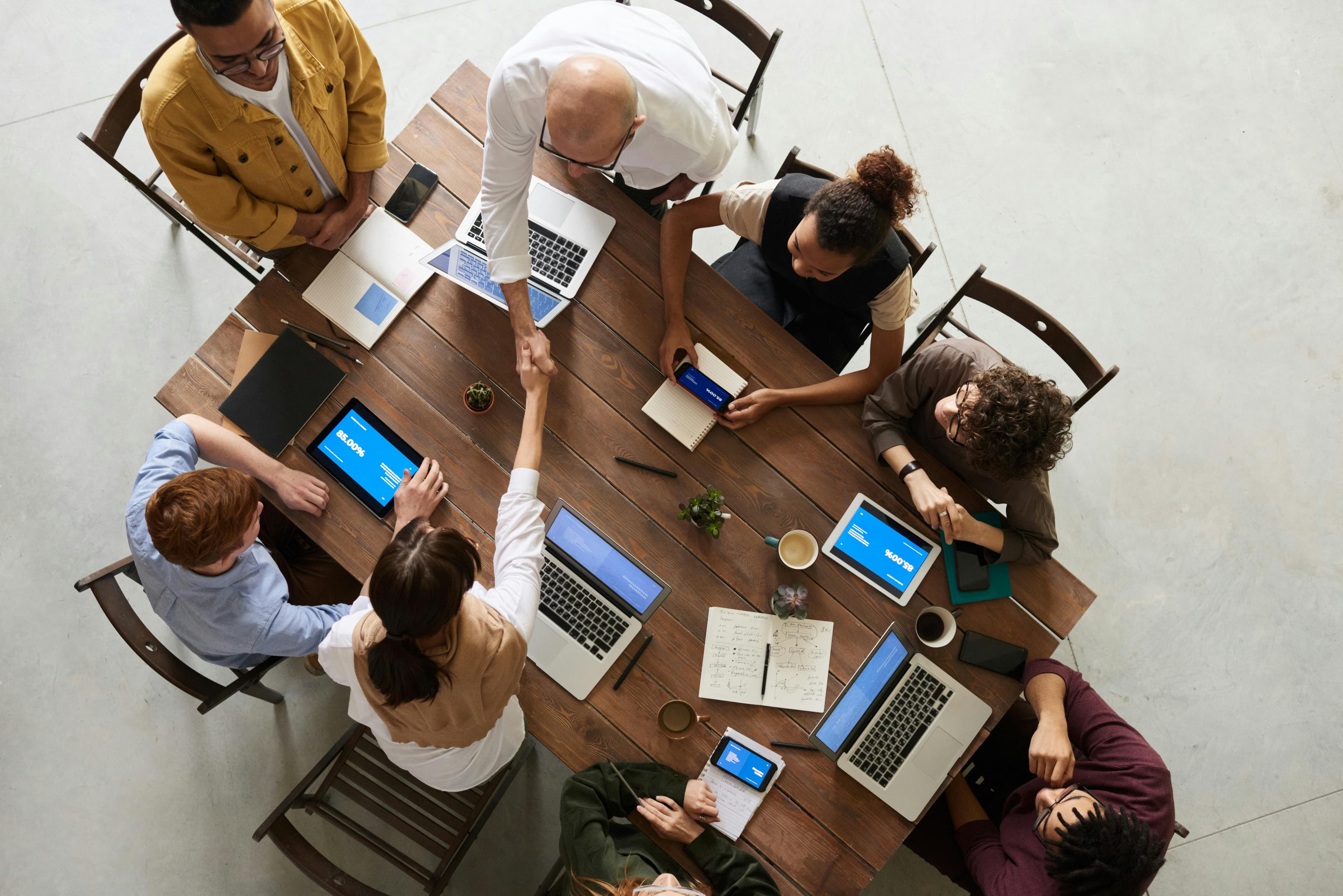 A group of people sitting at a table with various electronic devices, two of them shaking hands.